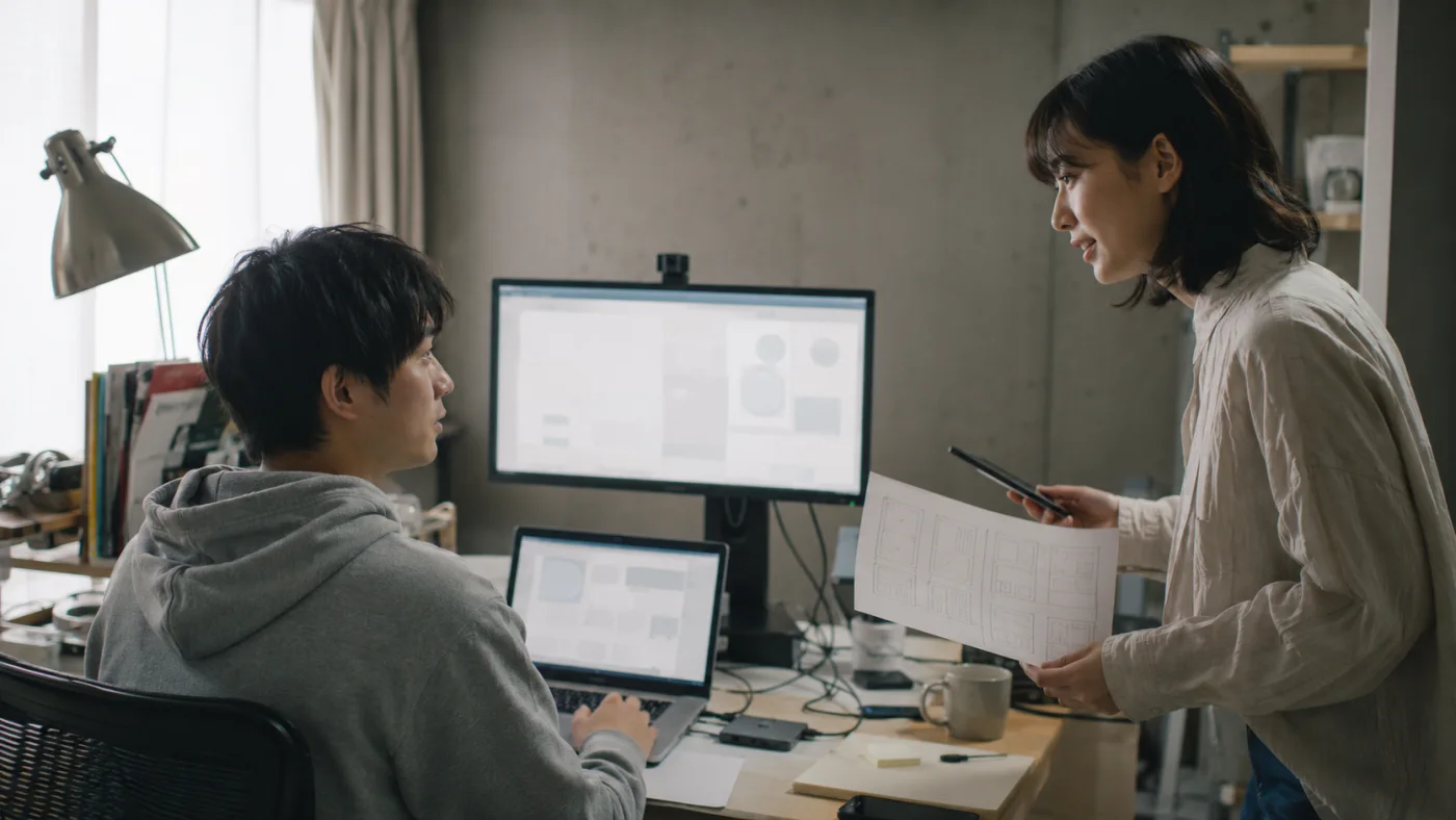 モニターの前でコードを書く知也と、改善案を持ち込むUIデザイナーの春菜