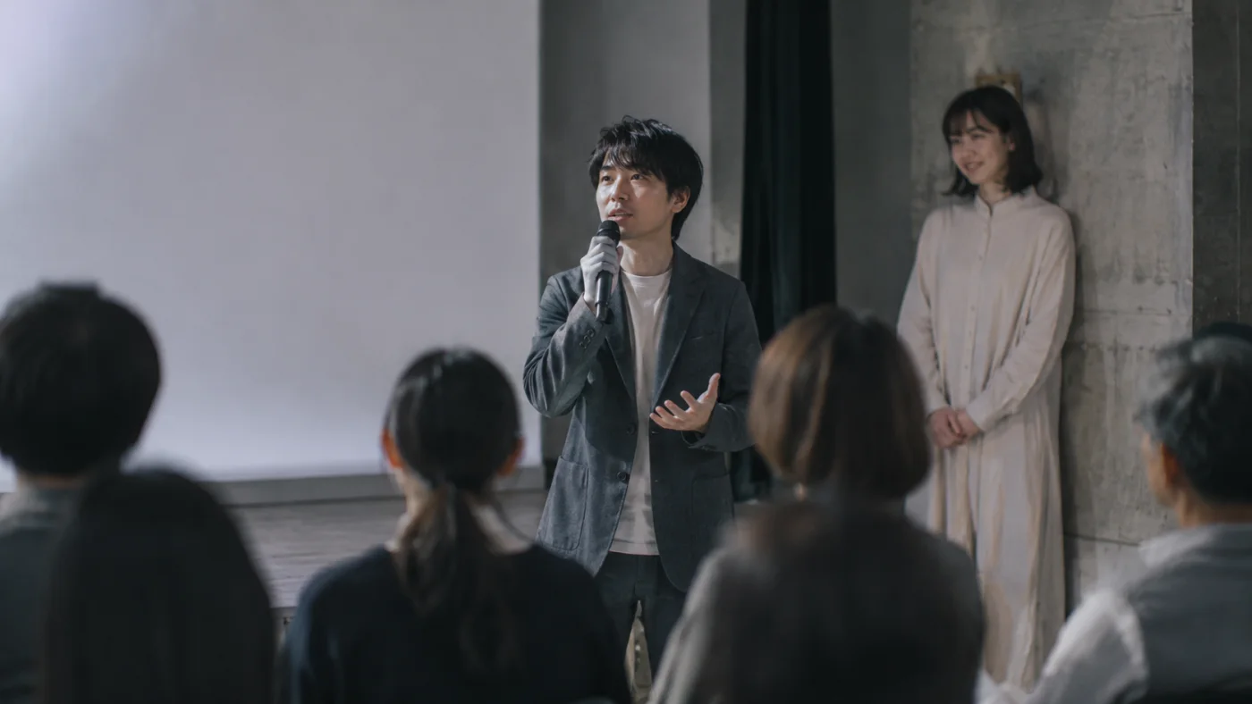 講演会場でマイクを手に語る知也と、後ろでにっこり聞く春菜